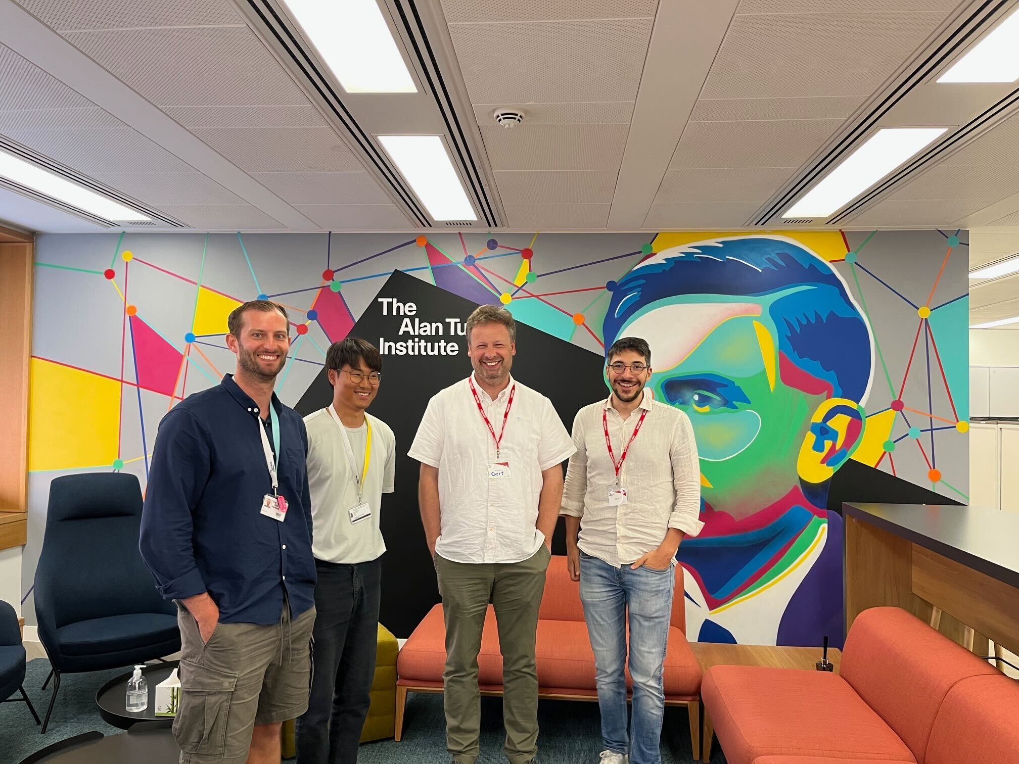 Four people stand between sofas in front of a sign reading 'The Alan Turing Institute'
